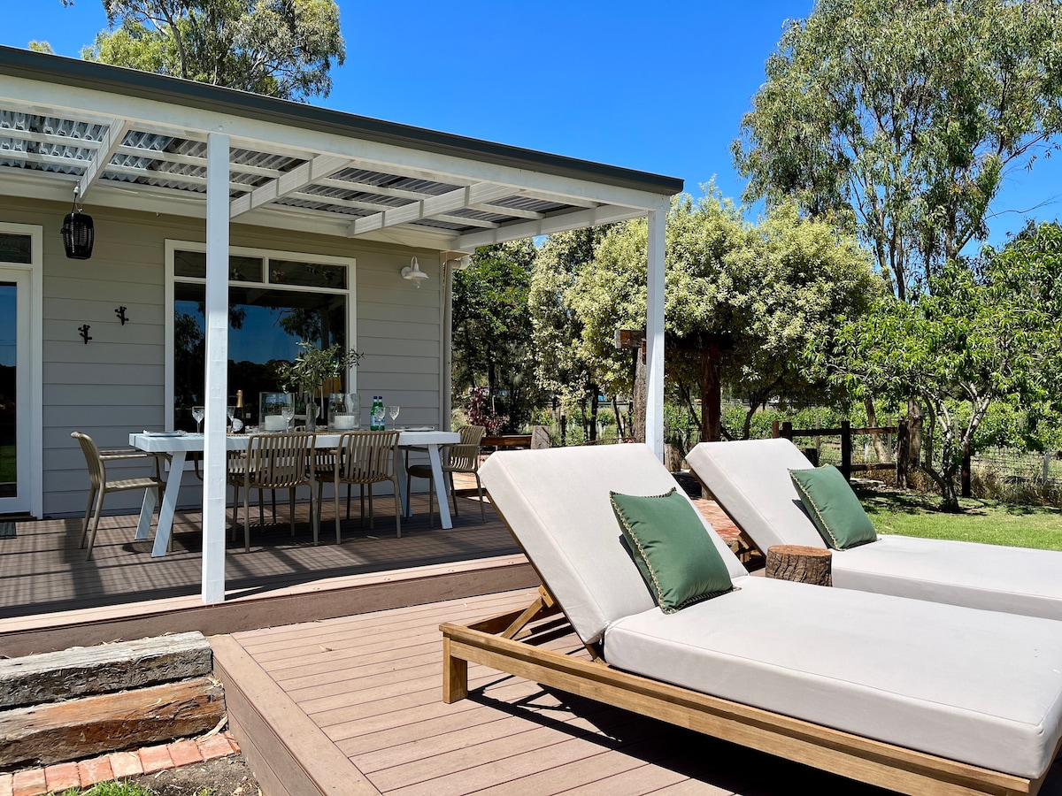 A sun deck is shown, featuring two lounge chairs with green cushions and a dining table with seating for six. The surrounding area includes lush greenery, and a shaded roof provides comfort from the sun.