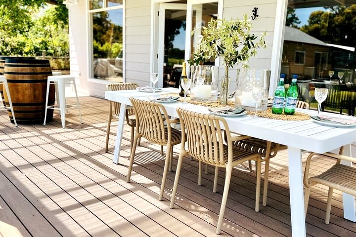 An outdoor dining area features a long white table set with elegant glassware and plates, surrounded by several beige chairs. A large barrel stands nearby, and lush greenery can be seen in the background, enhancing the inviting atmosphere of the sun deck.
