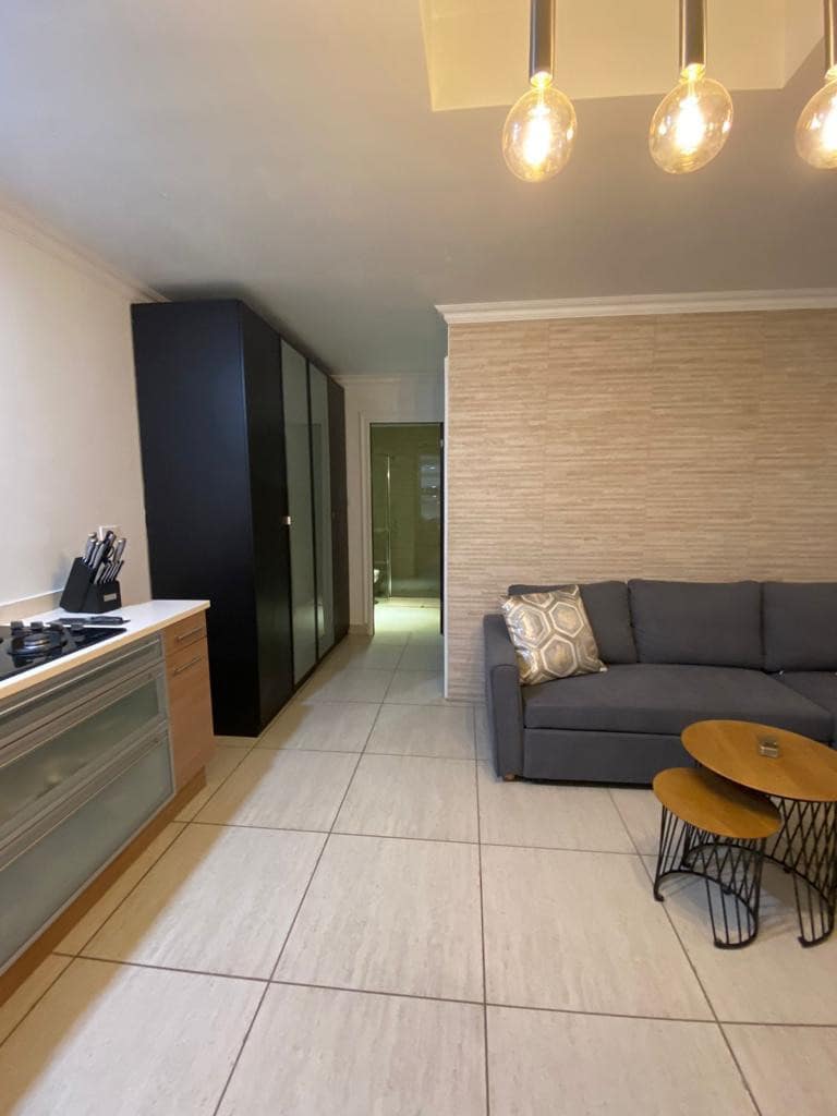 A modern kitchen corner features sleek cabinetry and is equipped with essential cooking utensils. A comfortable gray sofa is positioned nearby, paired with two round coffee tables. Soft pendant lighting illuminates the space, enhancing the clean lines of the tiled flooring.