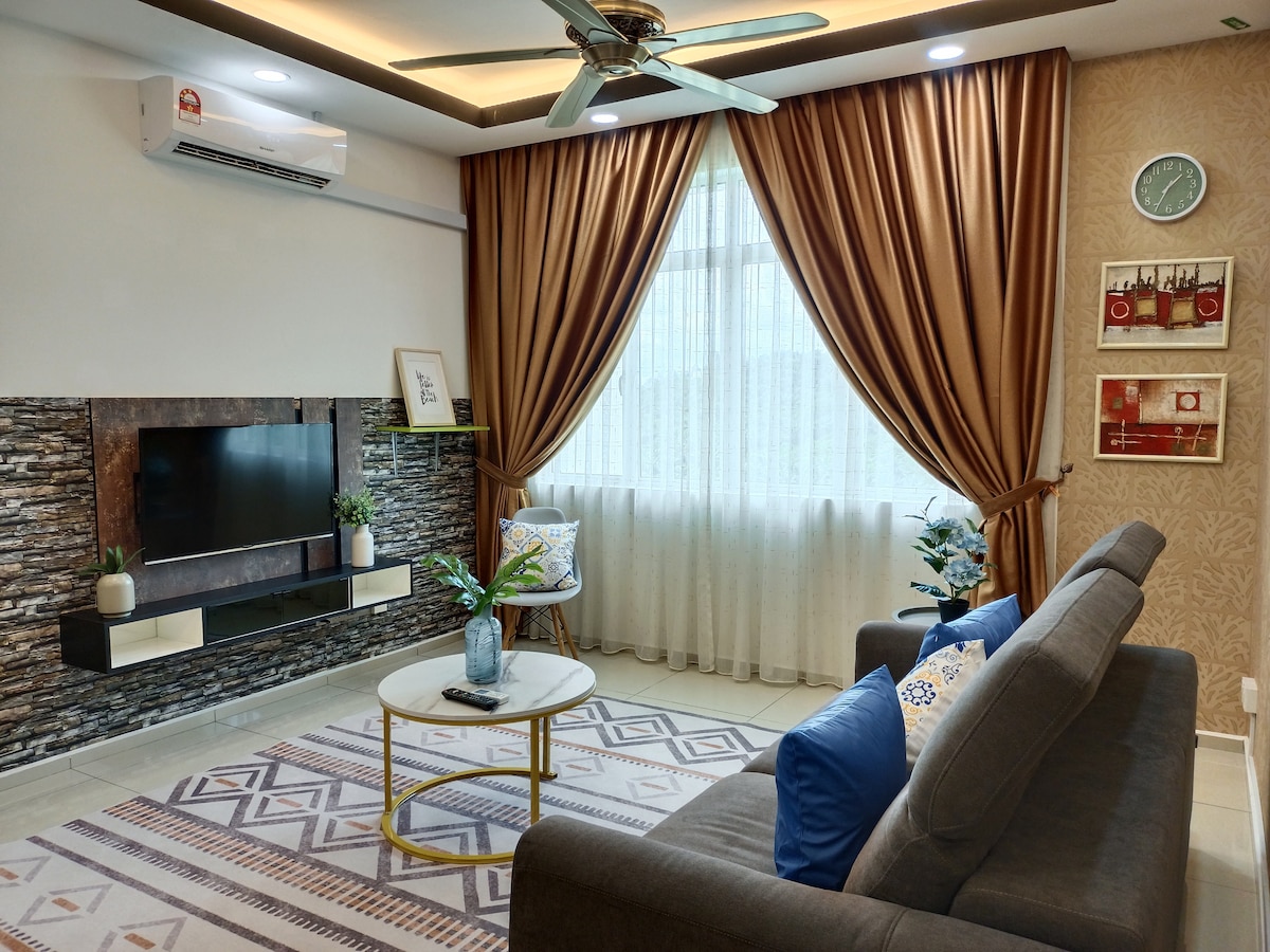 A living area is presented with a comfortable gray sofa, a round coffee table, and decorative cushions. Large windows covered with light curtains allow natural light to fill the space, while an air conditioning unit and wall-mounted television add to the functionality. A patterned rug complements the decor.