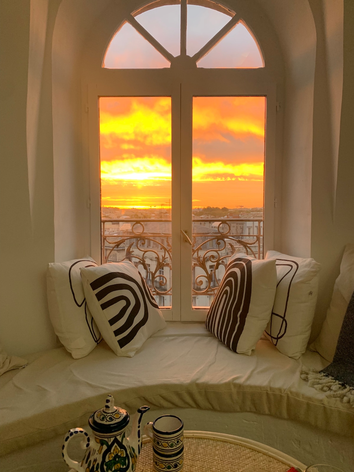A cozy nook is highlighted by a large arch window showcasing a vibrant sunset over Montpellier. Two patterned cushions rest on a soft seating area, while a decorated tea set awaits on a round table, enhancing the inviting feel of the space.
