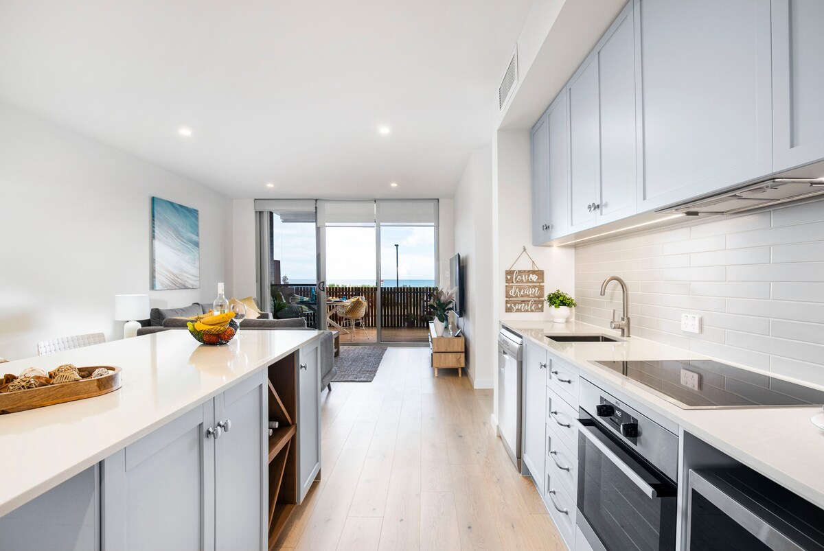 A modern kitchen is presented with light gray cabinetry and a spacious countertop. An open layout connects the kitchen to a living area, where natural light fills the space. A selection of fresh fruit is displayed on a wooden tray, adding a touch of freshness.