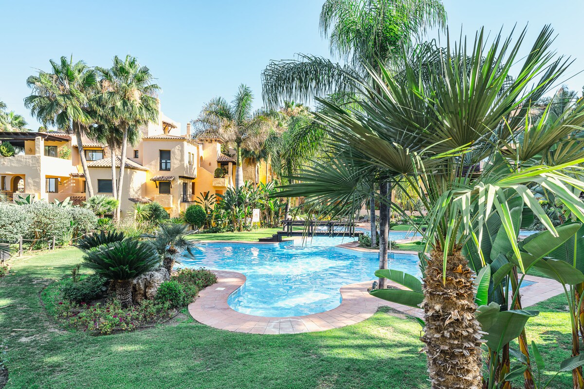A tranquil community pool is surrounded by manicured gardens and palm trees. The pool features a gentle curve and is partially shaded by the lush greenery. Nearby, sun loungers are positioned for relaxation, creating a peaceful outdoor space.