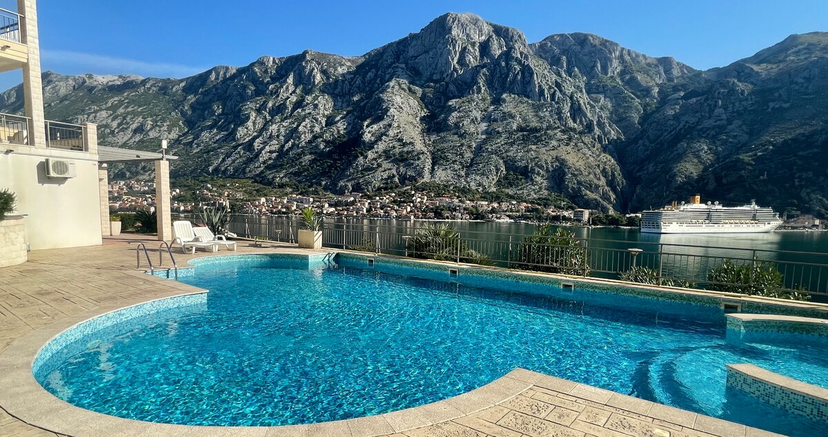 A large swimming pool glistens under the sun, framed by a stone patio. Towering mountains rise majestically in the background, with lush greenery visible along the shores. The serene bay reflects the clear sky, while a cruise ship can be spotted on the water's surface.
