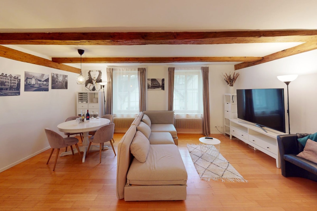 The living and dining area is designed with wooden beams overhead and features large windows that provide natural light. A round dining table with gray chairs is placed beside a cozy sectional sofa. A 65-inch smart TV is positioned on a white media console.