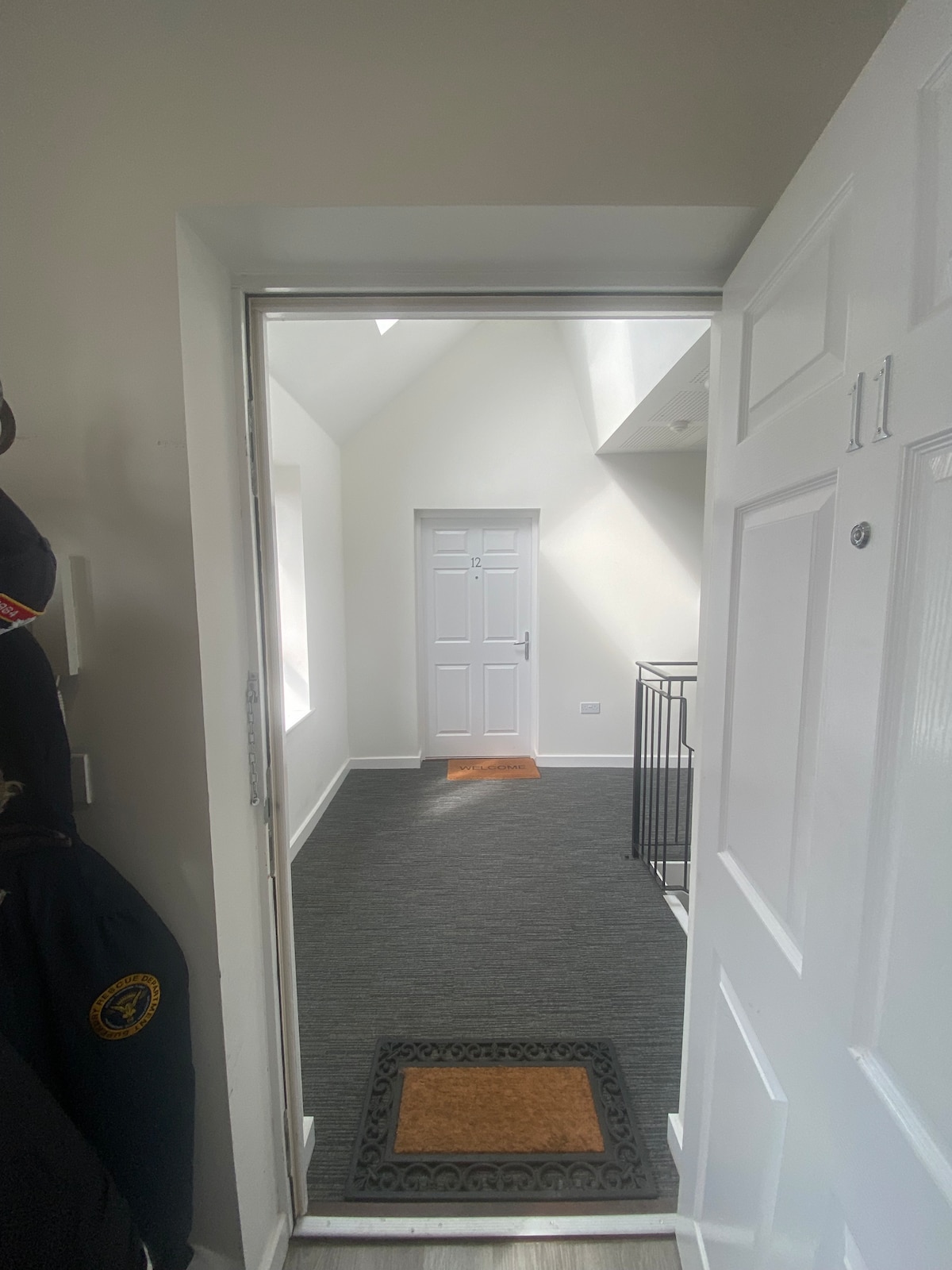 The entrance to the apartment is framed by a white door. A welcome mat lies at the doorstep, leading into a bright hallway adorned with neutral tones. The space features a door at the end and a staircase, enhancing the airy atmosphere.