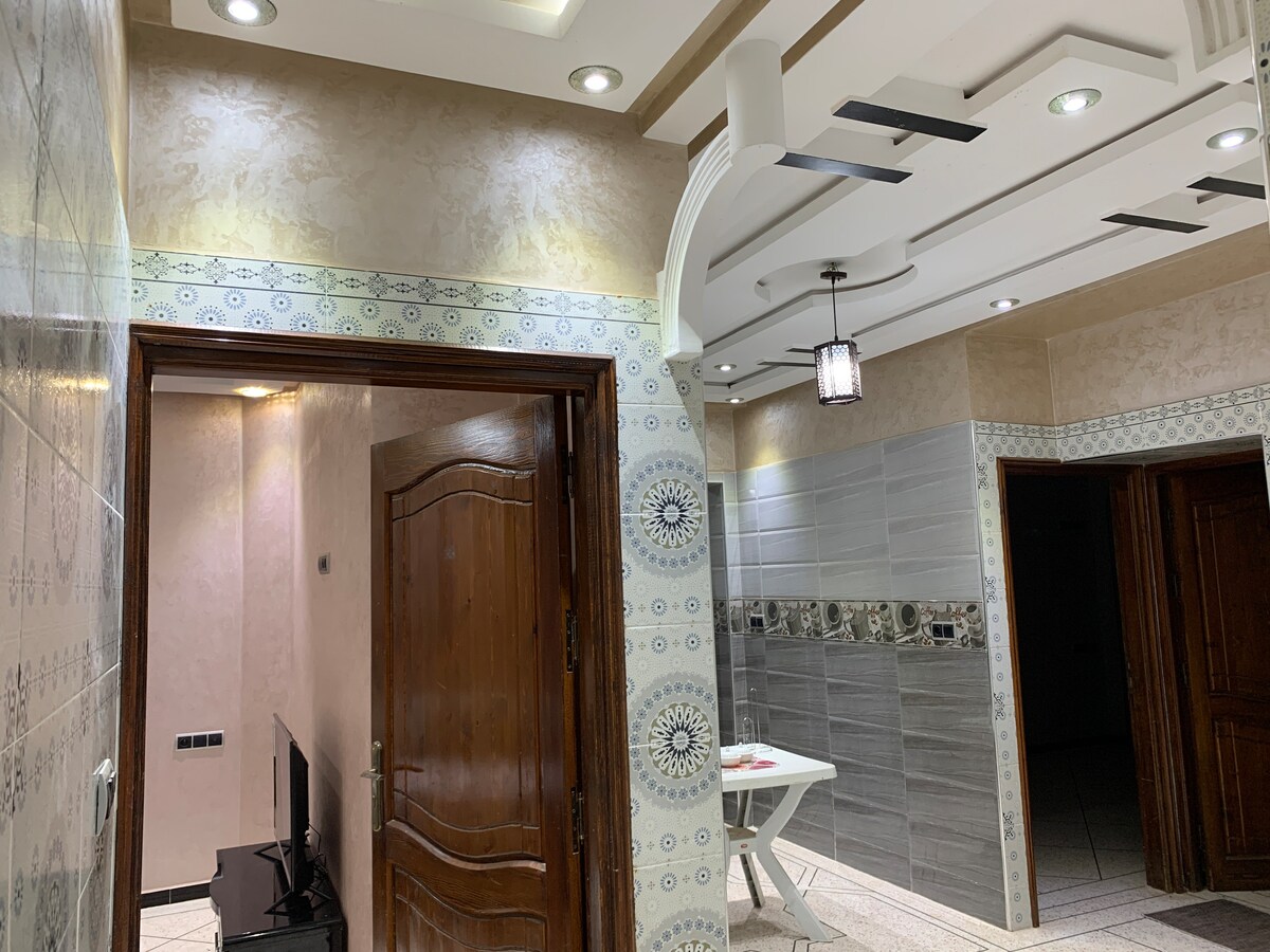 A well-lit hallway features decorative wall tiles and elegant molding. On one side, a wooden door leads to another room, while a small table is positioned within view. A television is visible just to the left, creating a welcoming feel.