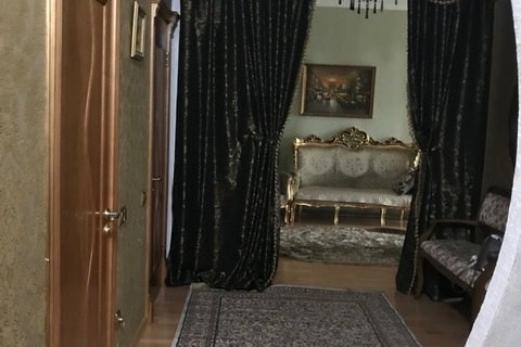 A hallway is framed by elegant, dark drapes, leading to a tastefully furnished sitting area. A vintage-style sofa with ornate detailing is positioned on a plush rug, complemented by a classic painting on the wall. Warm wood tones enhance the inviting ambiance.