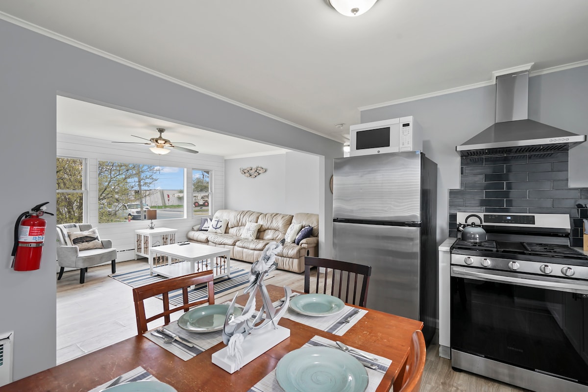 A dining area features a wooden table set for four, complemented by light blue dishes. Across the room, a spacious living area is visible, containing a beige sofa and large windows that allow natural light to fill the space.