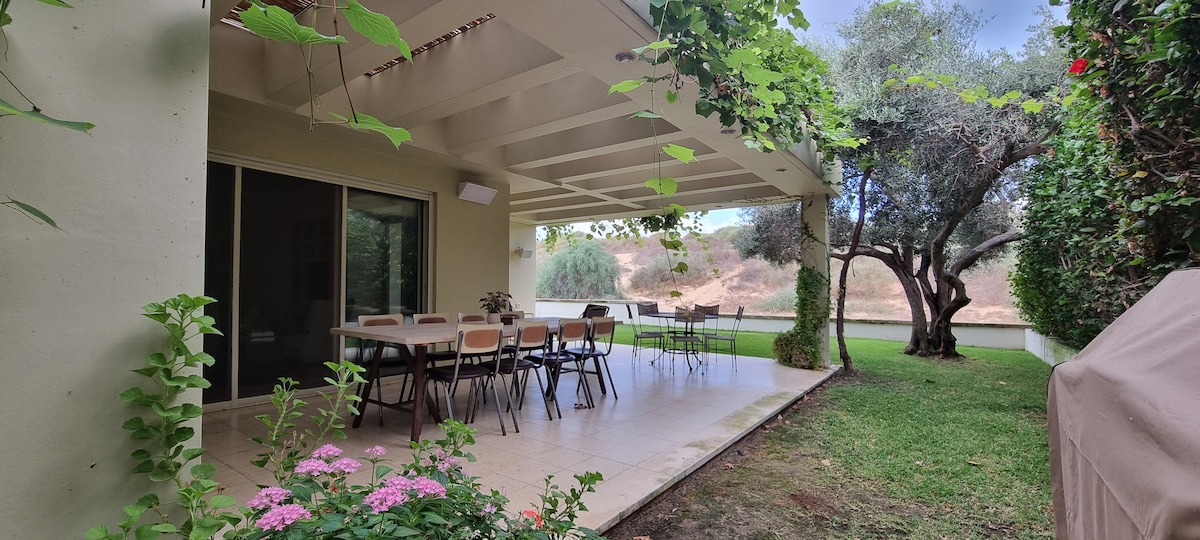 A covered outdoor patio features a spacious dining area with a large table and multiple chairs. Lush green vines adorn the wooden beams overhead. The grassy yard is bordered by trees, creating a serene atmosphere that extends towards a tranquil water view.