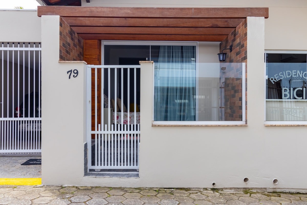 The entrance features a gated pathway leading to a welcoming porch. A combination of brick and wooden elements creates a modern façade. Large windows allow natural light to fill the space, while a light fixture adds a touch of elegance in the evening.