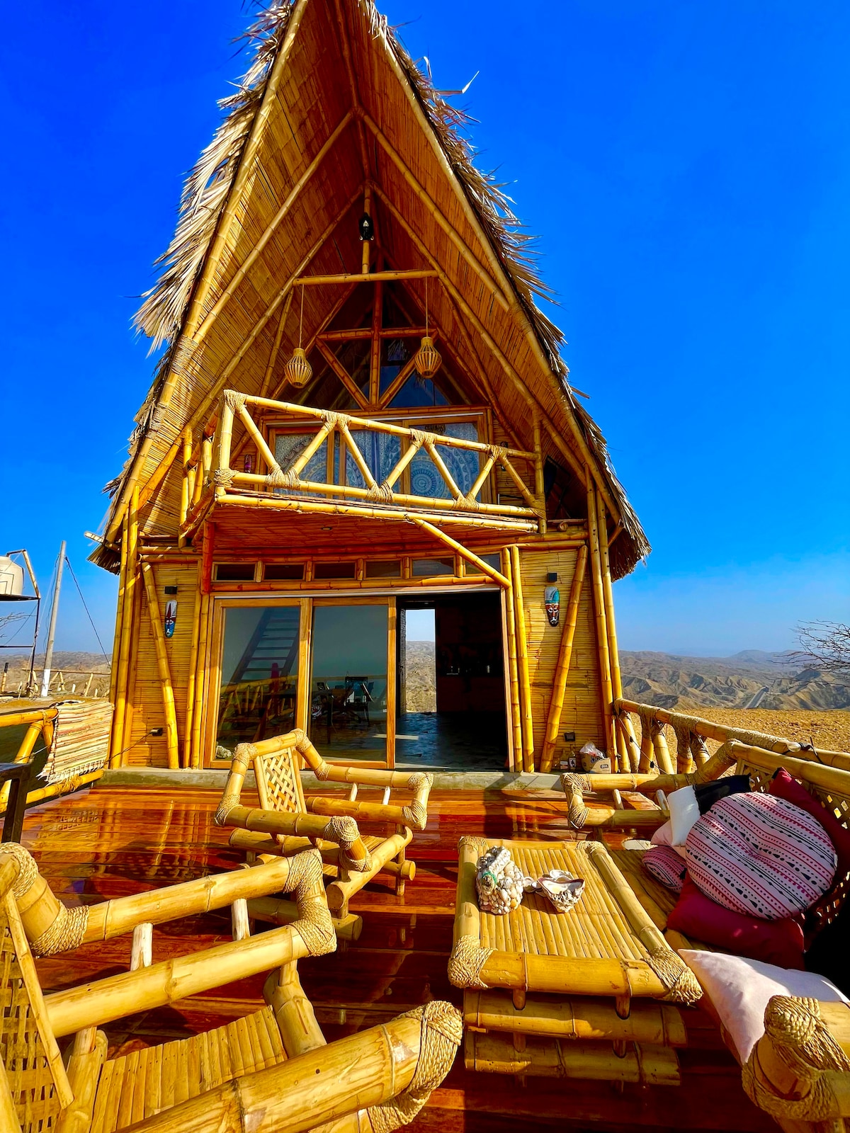 A bamboo and wood cabin is shown against a clear blue sky. The structure features a distinct peaked roof and large glass doors that provide a view of the surrounding landscape. Outdoor seating made from bamboo is arranged on the deck, inviting relaxation.