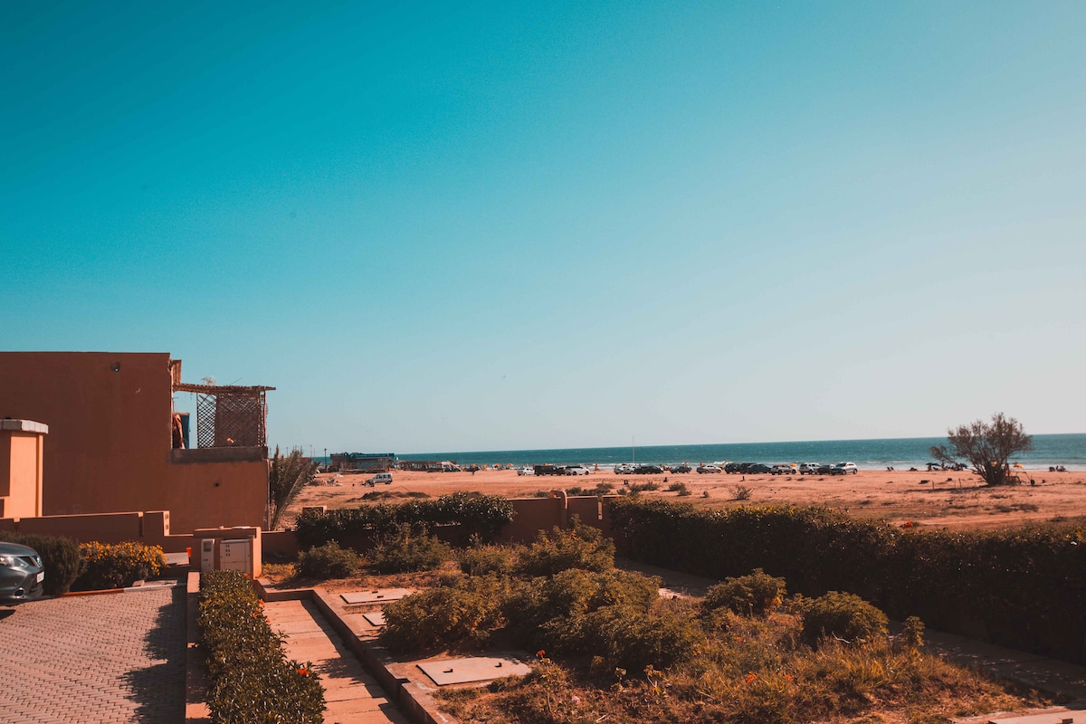A clear view of the beach and sea can be seen from the property. The sandy shoreline extends towards the horizon under a bright blue sky. Lush greenery is visible in the foreground, enhancing the natural setting. A few parked cars are located on a paved area nearby.