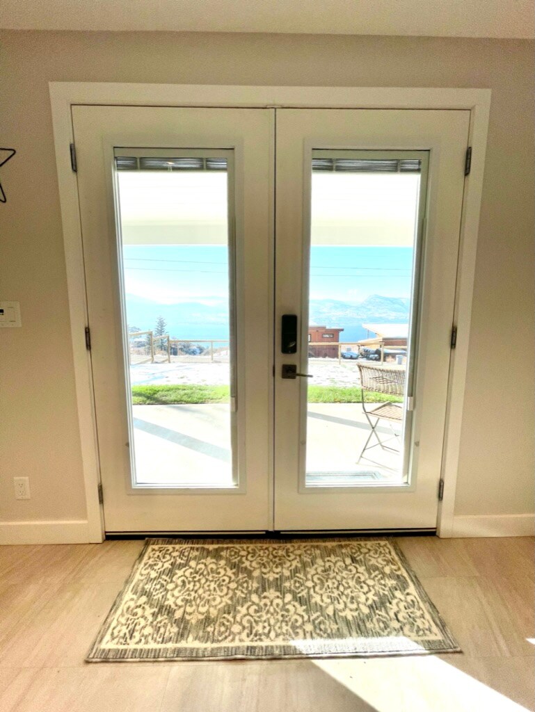 A pair of glass doors leads to an outdoor space, allowing views of the surrounding landscape. A doormat lies in front of the entrance, and a small outdoor bistro table with chairs is visible beyond the threshold.
