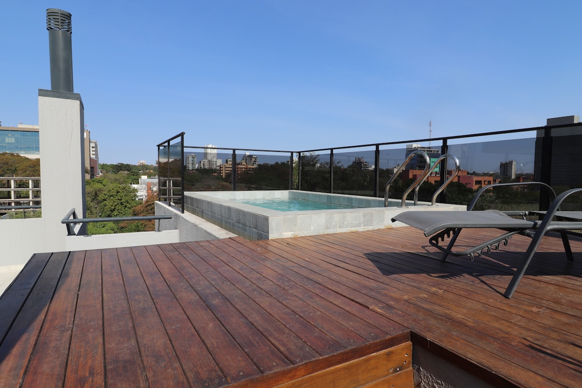 A rooftop area features a small dipping pool surrounded by a wooden deck. Metal lounge chairs are positioned nearby, offering a view of the surrounding area. The skyline and greenery are visible in the background under a clear blue sky.
