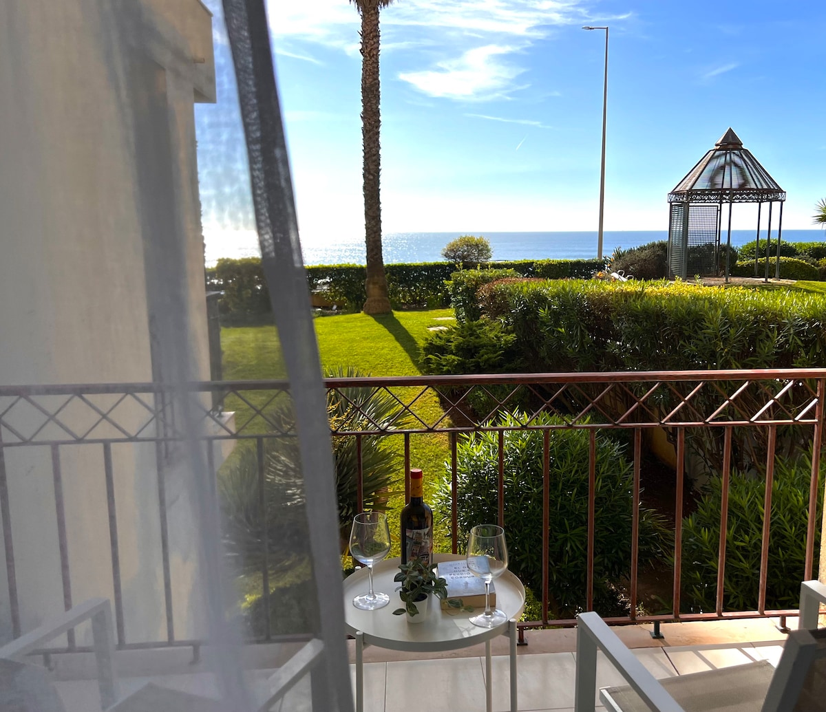 A serene balcony view showcases a table set for two with wine glasses and a bottle of wine. Lush greenery surrounds the area, and the ocean is visible in the distance under a clear blue sky.