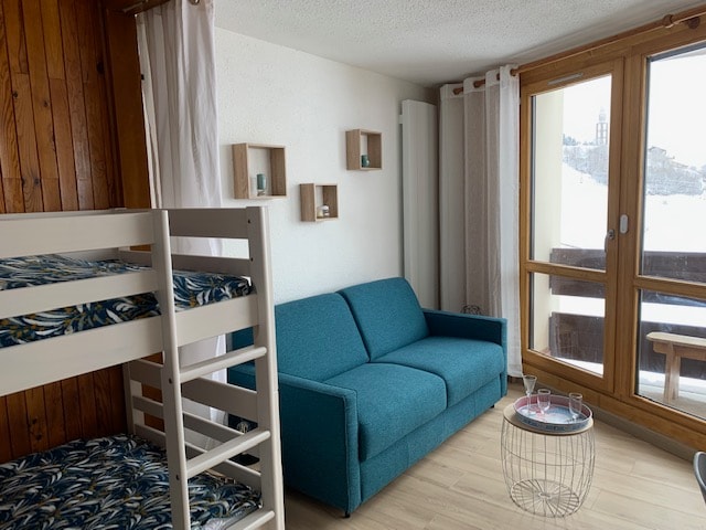 The studio features a bright living area with a turquoise sofa and a bunk bed. Natural light floods in through large windows, revealing a view of the surrounding mountains. A small, round table is positioned in front of the sofa.