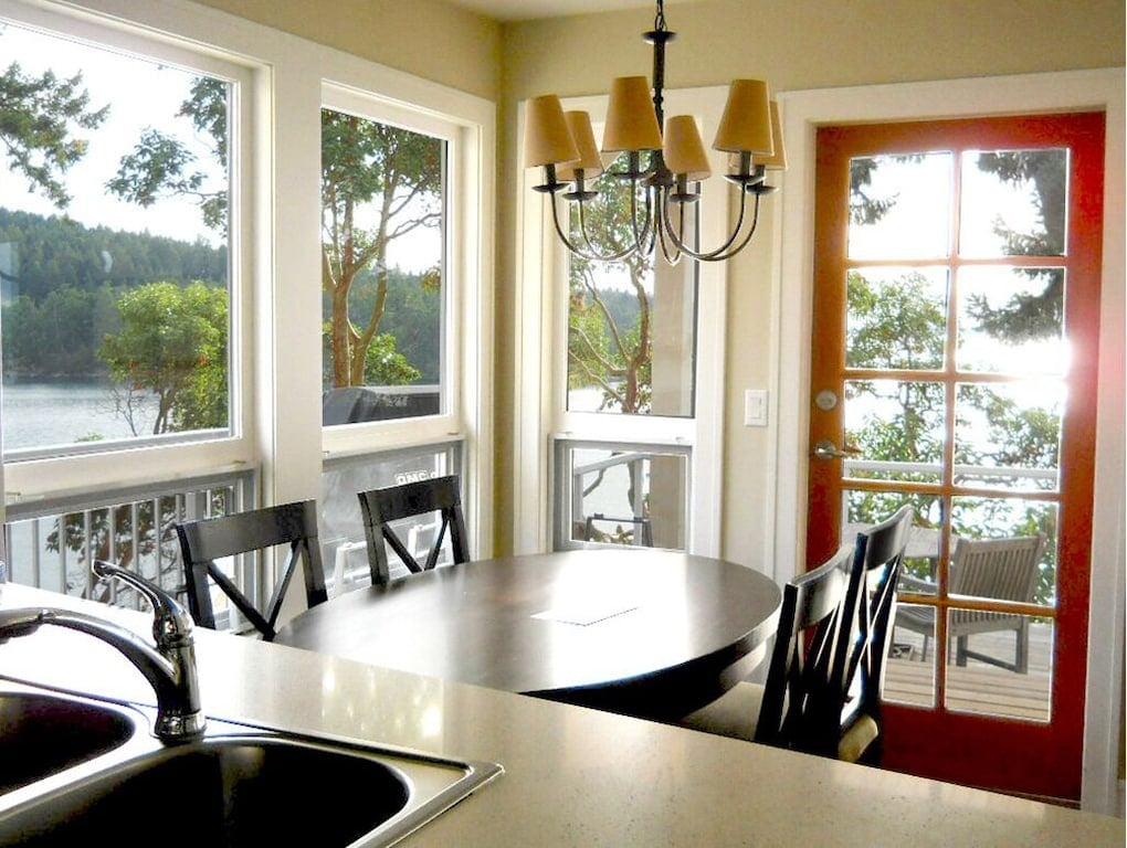 A sunlit dining area features a round table surrounded by black chairs, positioned under a chandelier. Large windows offer views of the surrounding trees and water, while a glass door leads to an outdoor deck, enhancing the connection to nature.