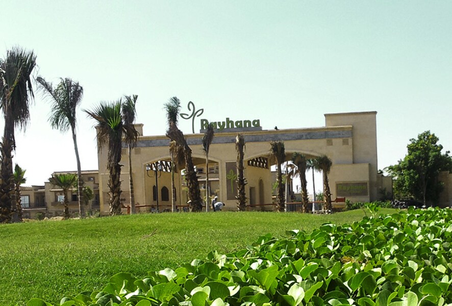 An elegant entrance is framed by tall palm trees and lush green landscaping. The building features warm-toned architecture, with the name 'Buhanga' prominently displayed above the entrance. A well-maintained lawn leads up to the entrance, enhancing the welcoming atmosphere.