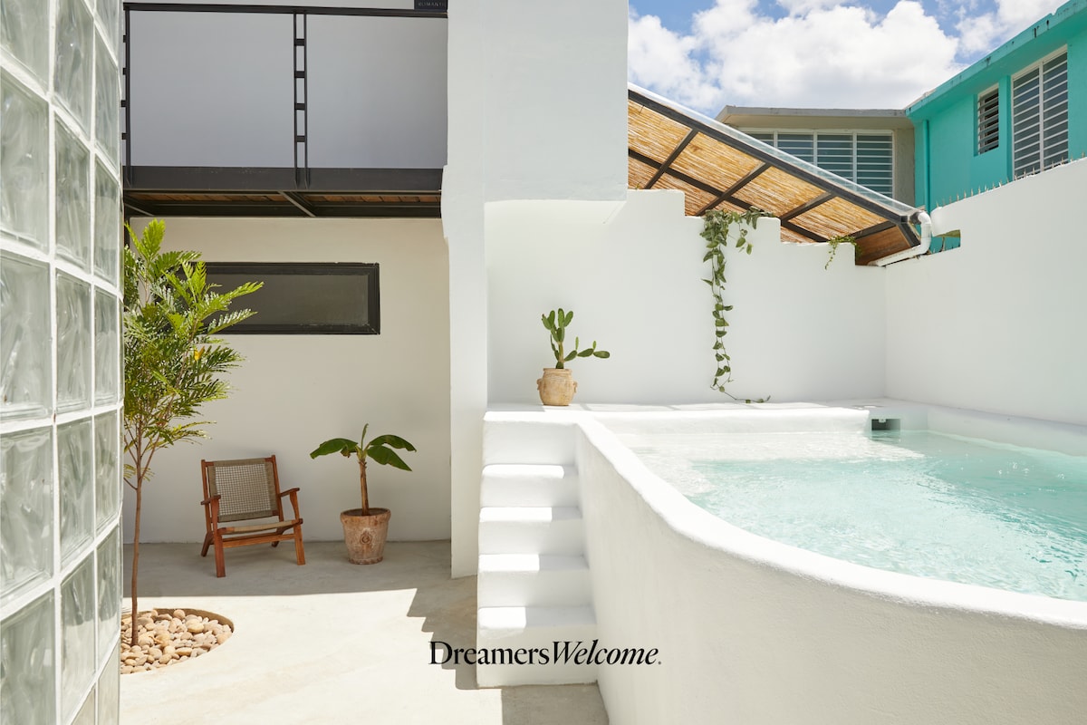 A serene outdoor area features a well-maintained pool bordered by a white wall and steps leading to a shaded terrace above. Lush green plants are placed strategically, and a wooden chair adds a touch of comfort to the space.