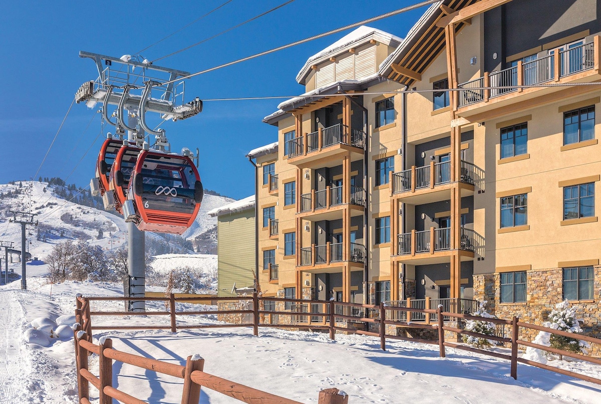 A ski lift runs alongside a modern building, featuring multiple balconies and large windows. Snow blankets the ground, creating a winter scene. The structure showcases a mix of earthy tones and stone accents, inviting guests to explore the surrounding slopes.