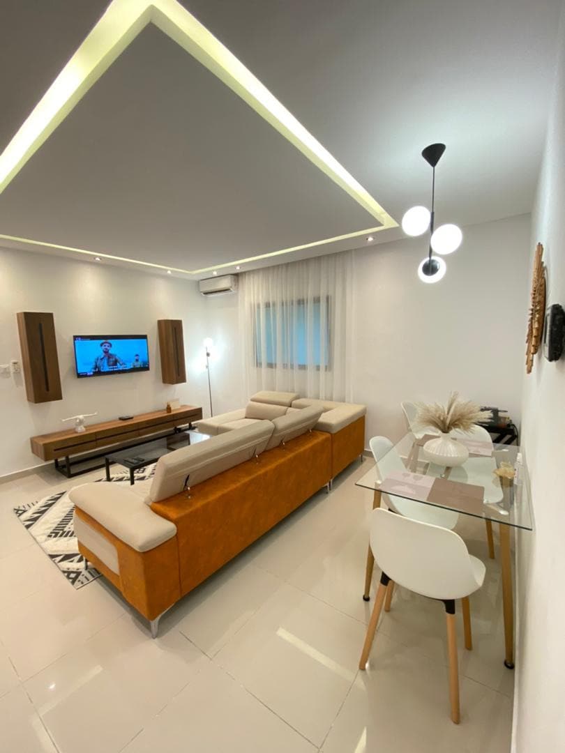 A modern living space is presented, featuring a comfortable sectional sofa and a wooden coffee table. A sleek television is mounted on the wall, while a dining table with white chairs is positioned nearby. Soft lighting highlights the serene atmosphere, complemented by a large window with sheer curtains.