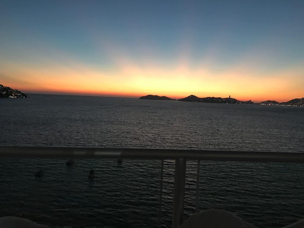 A serene sunset view over the bay is captured from a balcony. Soft hues of orange, pink, and purple blend in the sky, reflecting on the tranquil water. Silhouetted mountains are visible in the distance, creating a calming atmosphere.