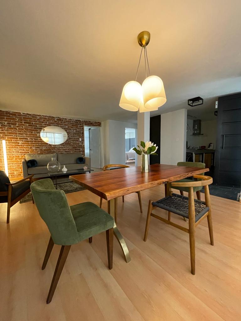 A modern dining area is highlighted by a wooden table surrounded by green and brown chairs. Soft lighting is provided by a distinct pendant fixture above. Exposed brick walls blend with a contemporary layout, creating a comfortable atmosphere for dining and gatherings.