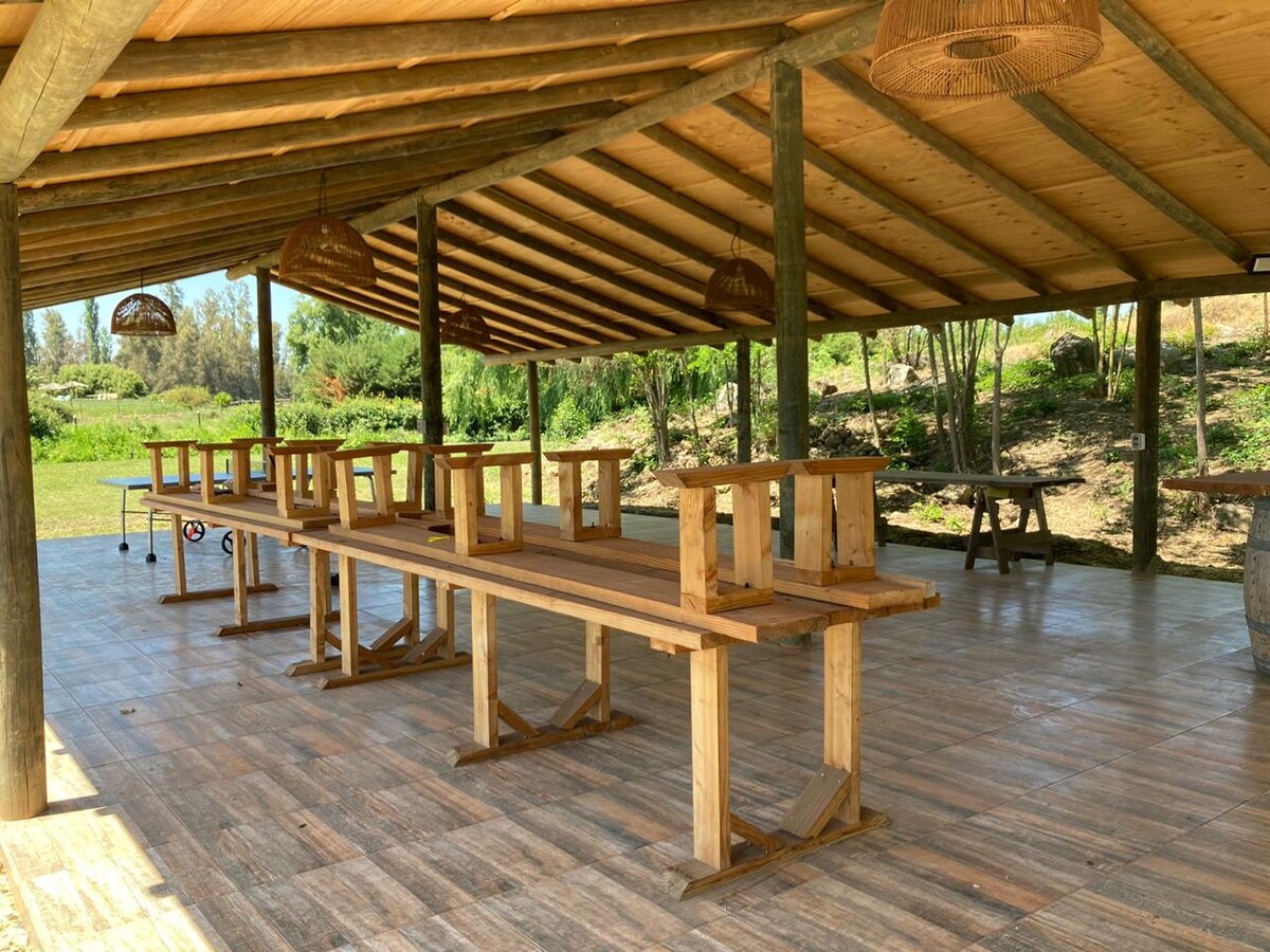 A spacious outdoor dining area features wooden tables and benches under a rustic roof. Natural lighting highlights the wooden flooring, while a serene landscape of greenery is visible beyond the open space. Enhanced by earthy tones, the structure encourages a communal atmosphere.