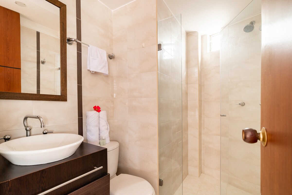 A modern bathroom features a glass-enclosed shower and a sleek vanity with a round sink. Light beige tiles adorn the walls, and a large mirror enhances the space. Fresh towels are neatly arranged, complemented by a single decorative flower.