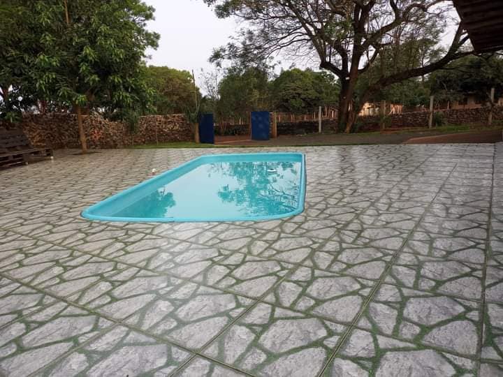 A rectangular pool with clear blue water is surrounded by a textured stone patio. Lush greenery and trees can be seen in the background, creating a serene outdoor space. The area is shaded, providing a cool retreat from the sun.