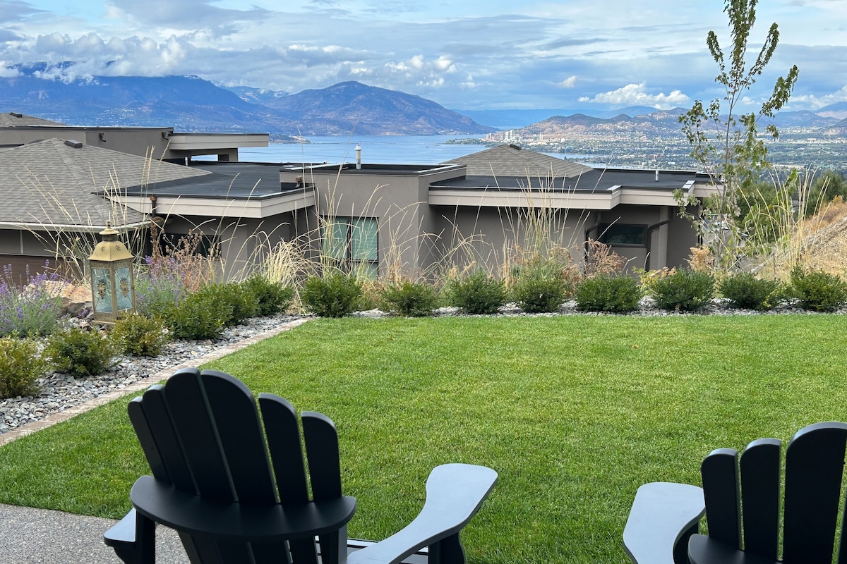 A scenic outdoor view features two Adirondack chairs positioned on a lush green lawn. The surrounding area is planted with low shrubs and ornamental grasses, framing a panoramic vista of the lake and distant mountains under a partly cloudy sky.