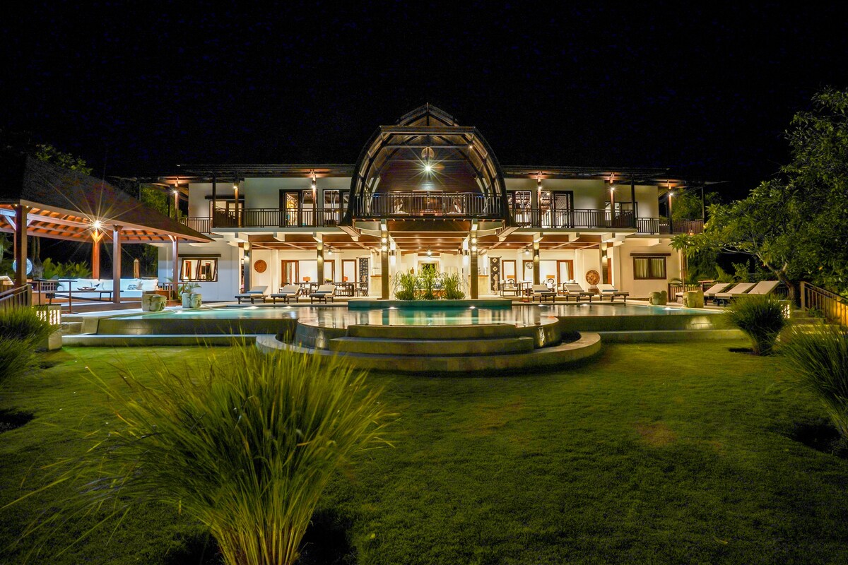 The villa's exterior is illuminated at night, highlighting the spacious pool with a serene ambiance. The lush green lawn frames the pool area, while the architecture features large balconies and multiple doors that create an inviting atmosphere. A gazebo is visible to the left.