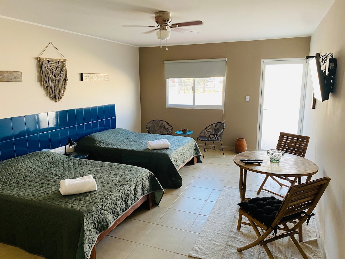 A comfortable suite is equipped with two spacious beds, each adorned with neatly folded towels. A small table and chairs occupy one corner, while natural light filters through the large window next to the door leading outside.