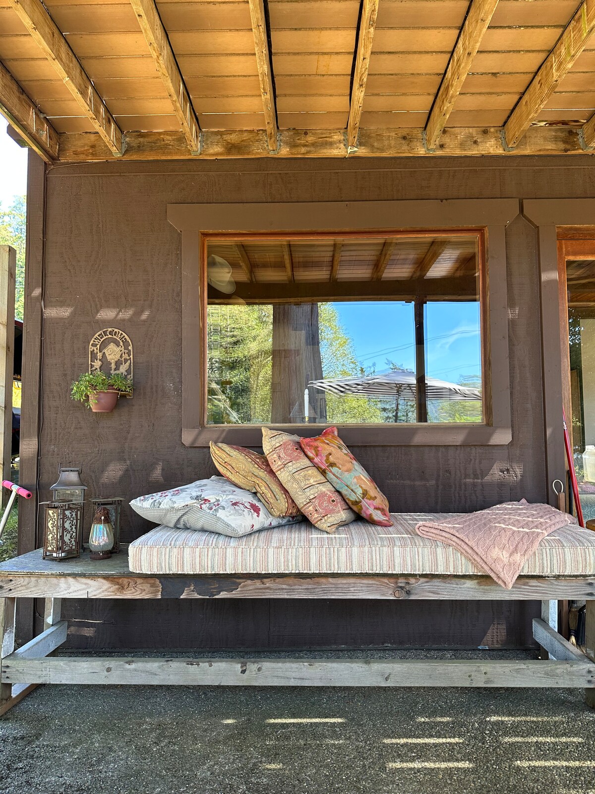 A cozy outdoor seating area is featured, with a cushioned bench adorned with patterned pillows. A lantern and flower pot are placed nearby, complementing the natural light that enters through the window above. The wooden structure creates a warm and inviting atmosphere.