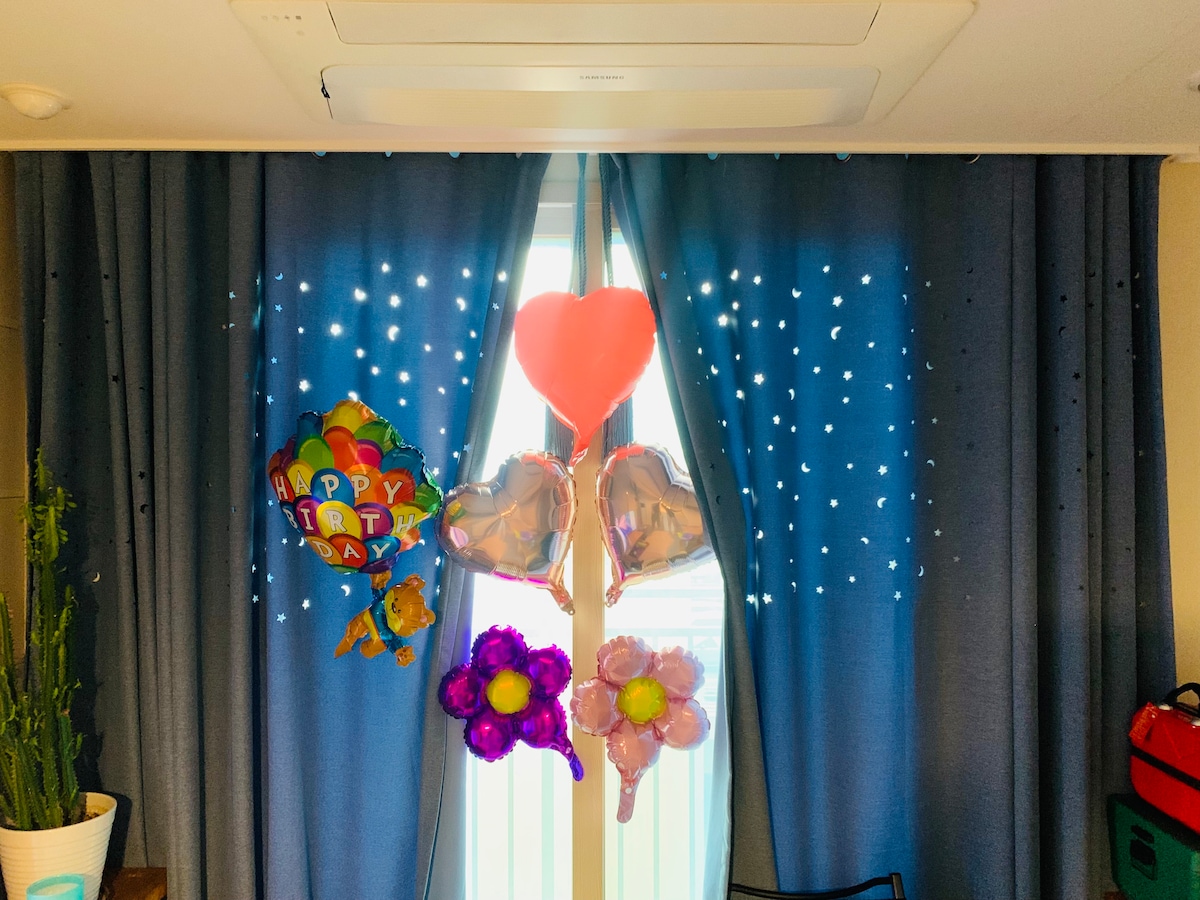 A window is adorned with blue curtains enhanced by lights, creating a backdrop for a collection of balloons. Brightly colored balloons include a birthday greeting, hearts, and flowers, adding a festive touch to the space.