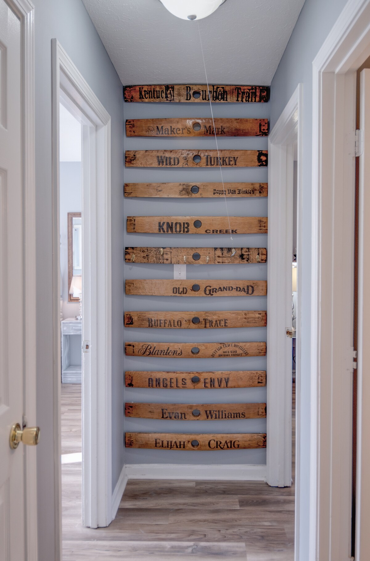 A narrow hallway is adorned with vintage bourbon barrel heads, displaying various brand names arranged vertically. The neutral-colored walls and flooring provide a subtle backdrop, leading towards a visible doorway at the end of the hall, inviting guests into the living space.