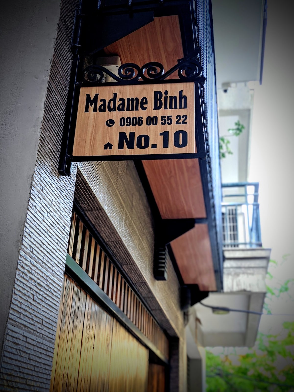 A wooden sign displays the name 'Madame Binh' alongside the contact number and the address 'No.10'. The sign's design features a blend of modern and traditional elements, hanging from an ornate metal bracket. Natural light illuminates the facade, giving a welcoming appearance.