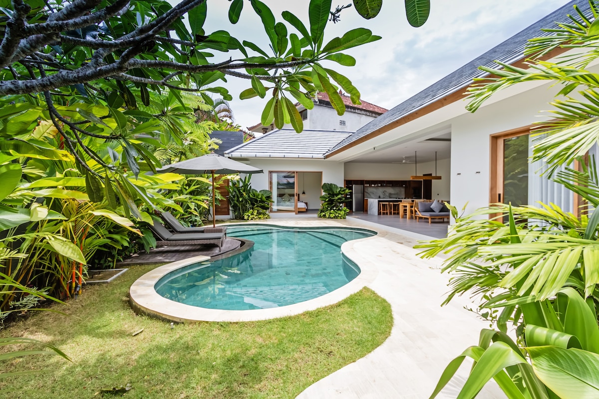 The image captures a serene outdoor area featuring a private swimming pool surrounded by lush tropical vegetation. Sun loungers and a large umbrella are positioned on the sun deck, while the open-plan kitchen and living space are visible in the background, enhancing the inviting ambiance.