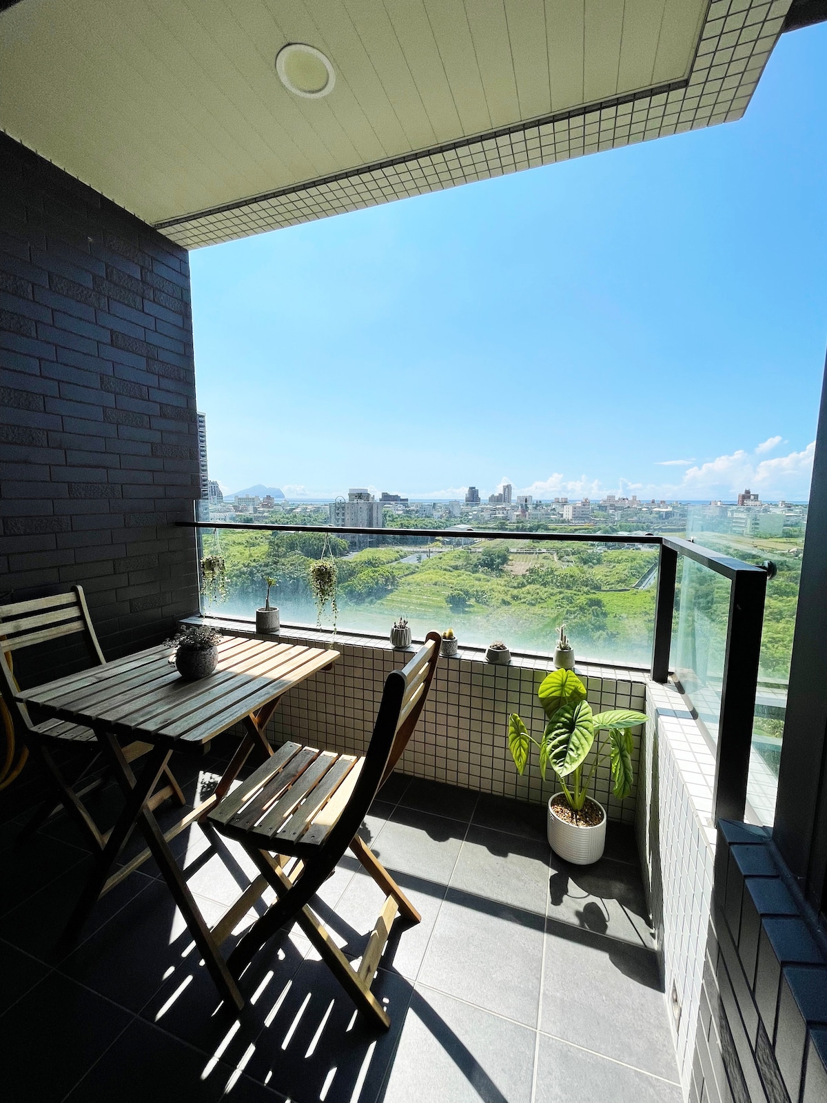 A balcony space features a small table and two wooden chairs, complemented by potted plants. The view showcases a vast landscape with greenery and distant city buildings under a clear blue sky, creating a serene outdoor setting.