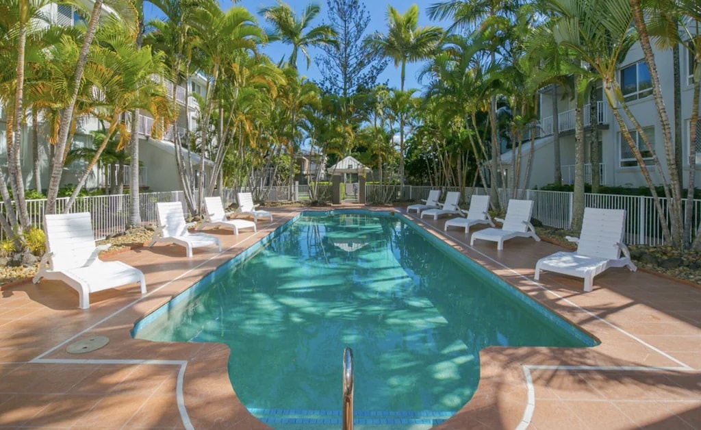 A serene pool area is framed by lush palm trees, creating a relaxing environment. Eight sun loungers are positioned around the pool, inviting guests to unwind. The clear water reflects the bright blue sky, complementing the tranquil setting.