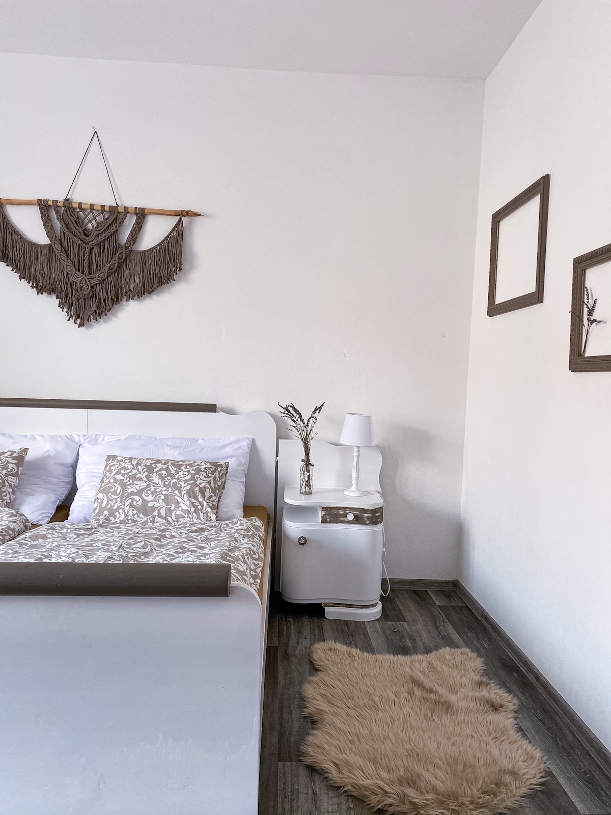 A serene bedroom setting features a spacious bed adorned with neutral-toned bedding and decorative pillows. A bedside table holds a lamp and decorative elements. Natural light may filter through, highlighting the textured wall hanging and soft rug positioned beside the bed.