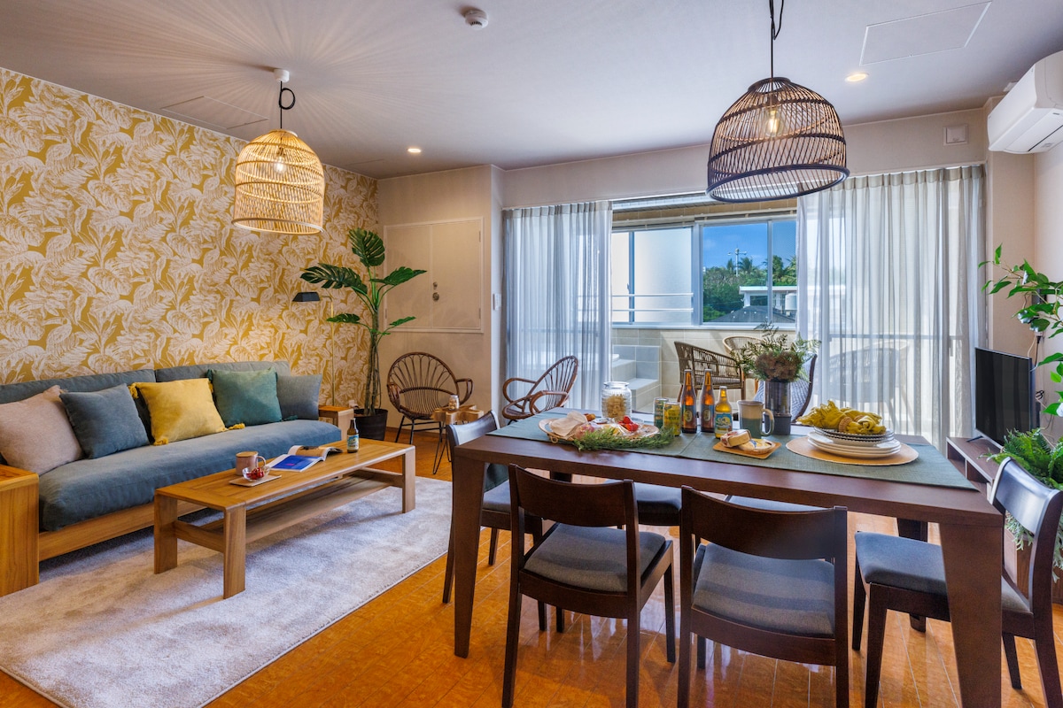 The living and dining area features a spacious seating arrangement with a sofa and wooden table. Natural light flows through large windows, highlighting the tropical-themed wall. A dining table is set for a meal, complemented by decorative plants and woven light fixtures.