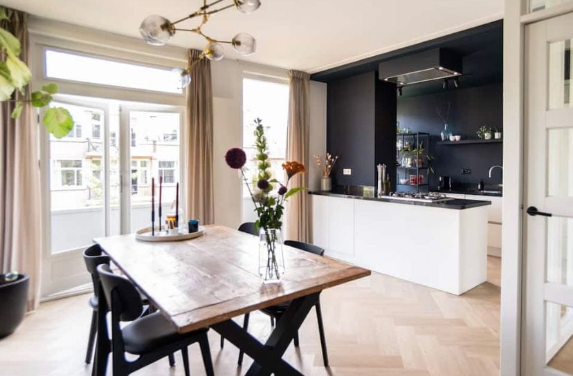 A modern kitchen with a large island features sleek black cabinetry and stainless steel appliances. A wooden dining table is surrounded by black chairs, complemented by a vase of fresh flowers. Natural light is invited in through large glass doors leading to a balcony.