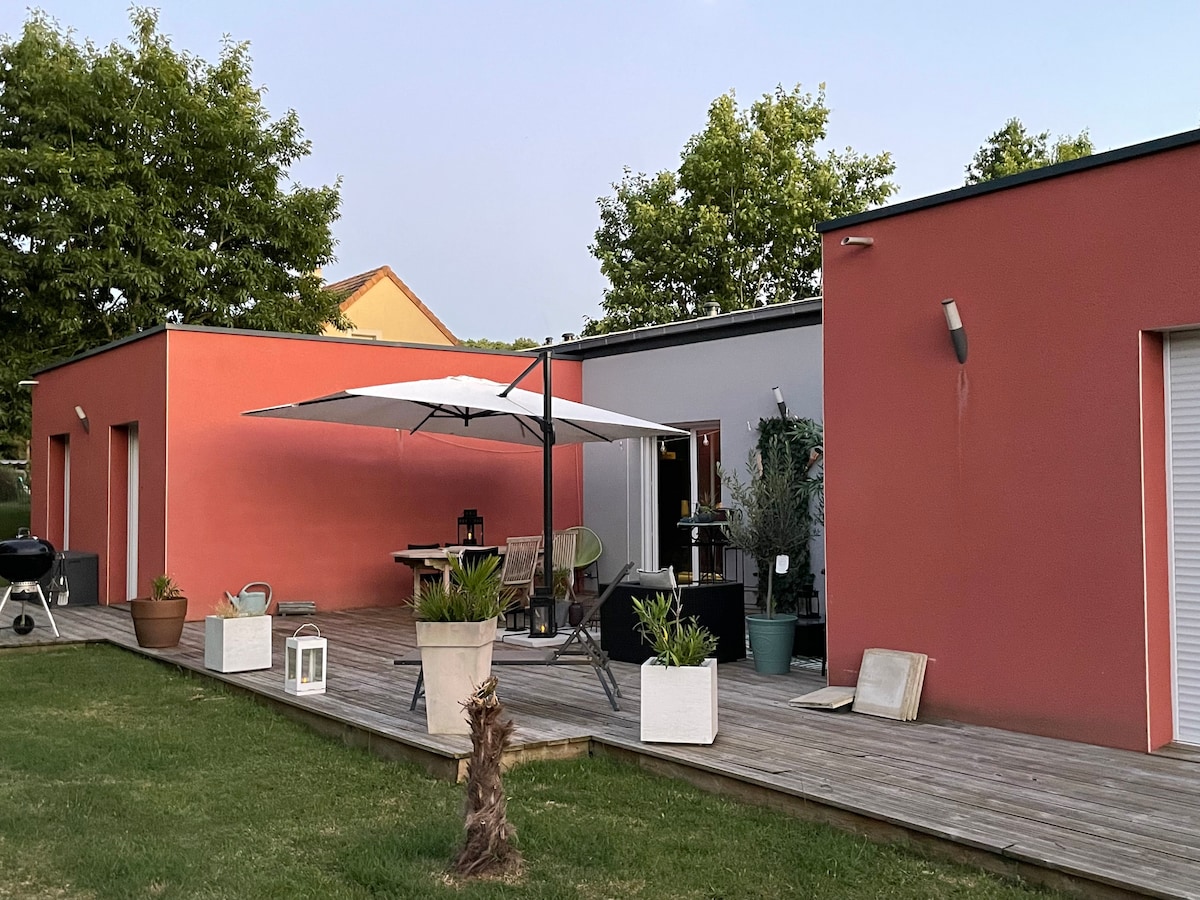 A modern exterior features a spacious wooden deck surrounded by well-maintained greenery. An umbrella provides shade over a dining area, with an outdoor seating arrangement visible. The house showcases a combination of red and neutral walls, reflecting a contemporary design.