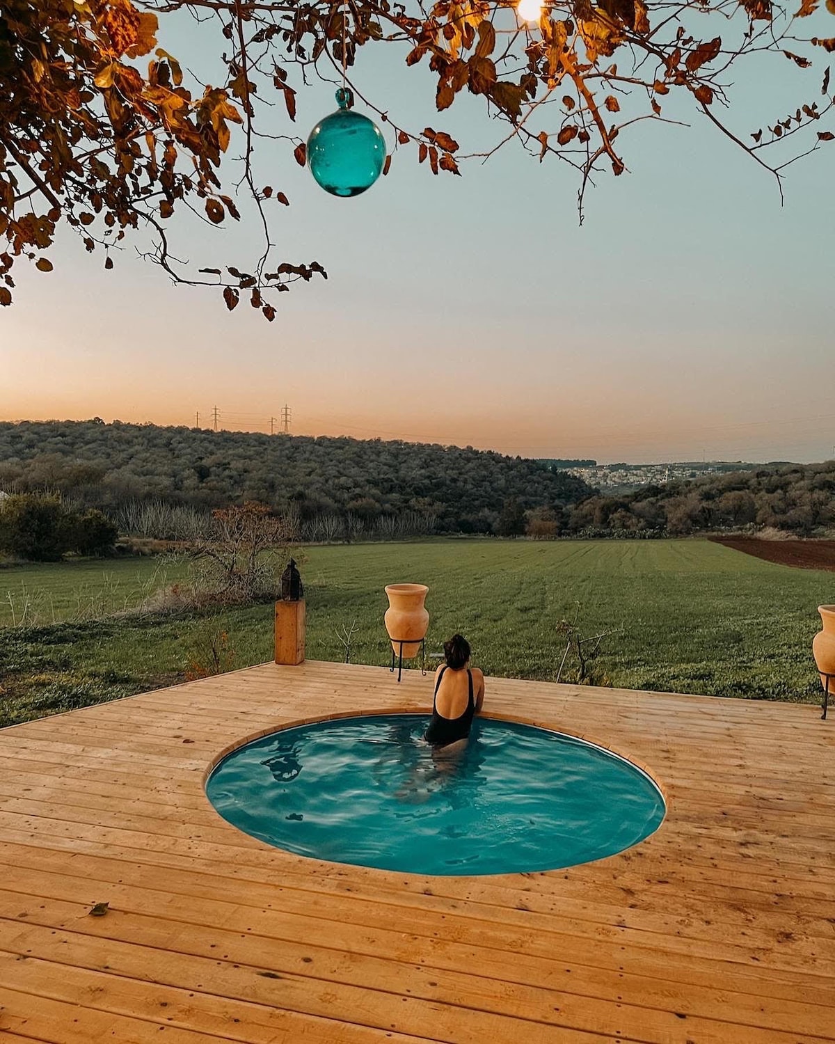 A round wooden deck supports a private hot tub, gently illuminated by soft lighting from above. A person is seen relaxing in the water, with a backdrop of rolling green fields and a soft sunset sky, providing a serene and tranquil atmosphere.