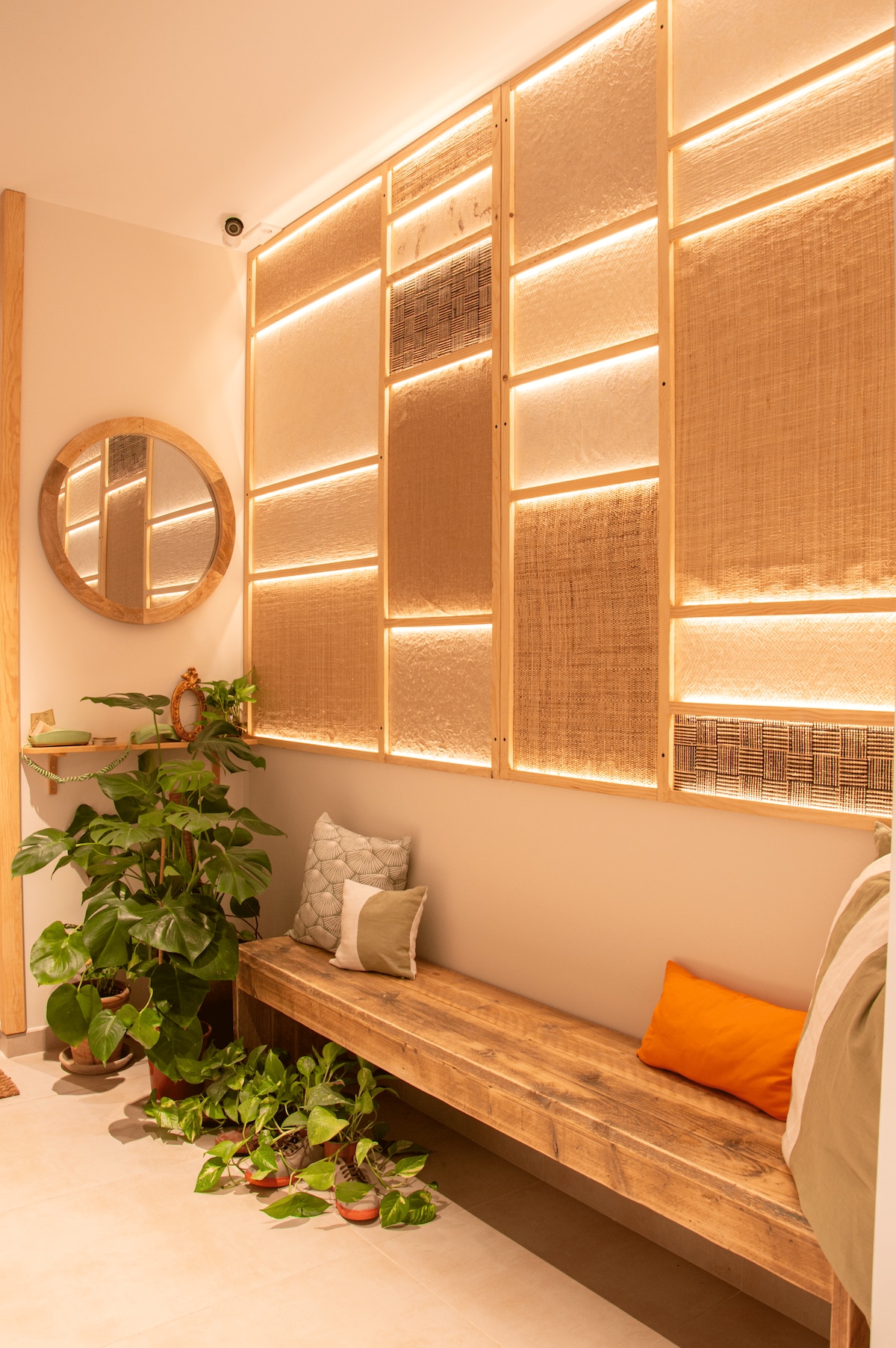 A welcoming entryway features a wooden bench adorned with decorative pillows and a variety of green plants. A circular mirror reflects softly lit wall panels, creating a cozy ambiance. Natural light enhances the warmth of the space, inviting guests to relax upon arrival.