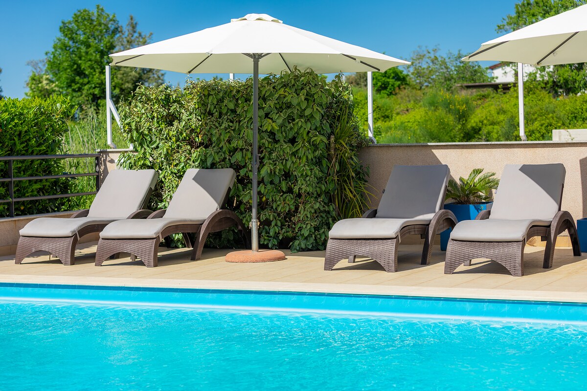 Four lounge chairs are arranged beside a swimming pool, positioned under white umbrellas. Lush greenery is visible in the background, providing a serene setting. The deck features light-colored wooden flooring, enhancing the outdoor relaxation area.