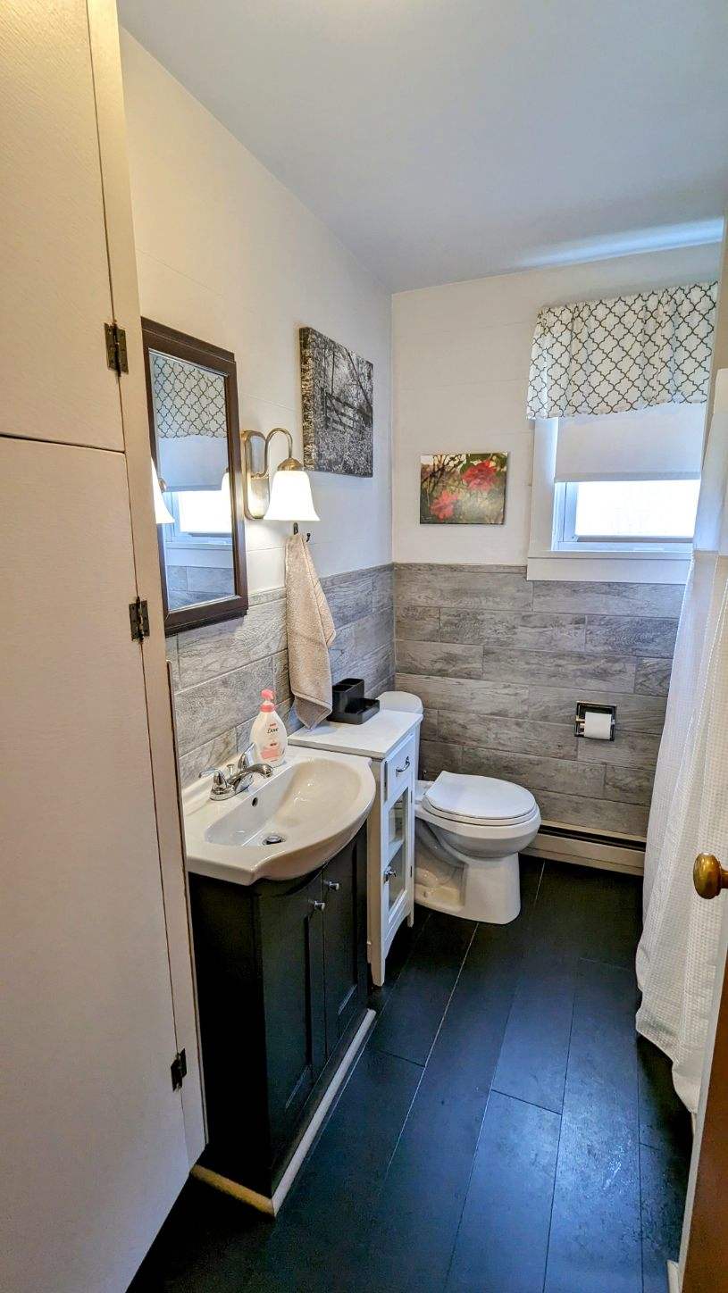 A compact bathroom features a dark vanity with a sink and cabinet beneath. A white toilet stands against the side wall. Natural light enters through a window adorned with a patterned valance. Art pieces are displayed on the walls, adding charm to the space.