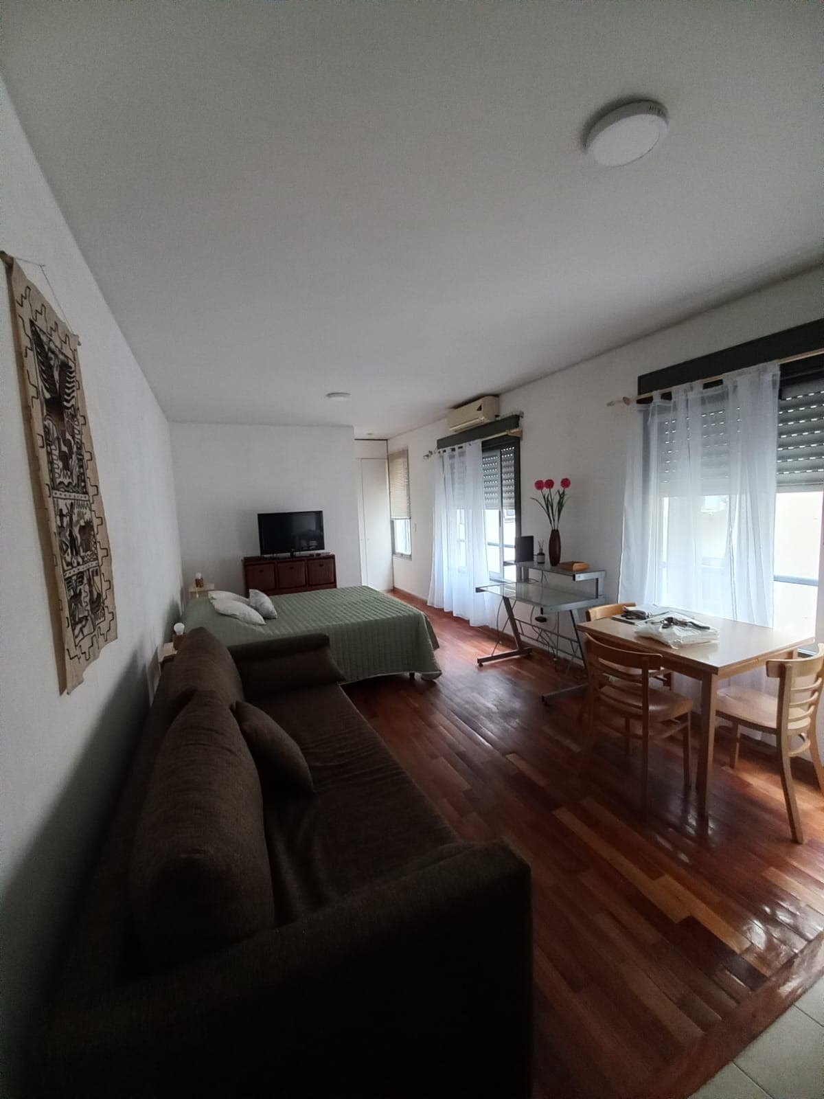 A comfortable living area is furnished with a dark brown sofa and a single bed draped in a green cover. A television is placed on a wooden unit, while a dining table with two chairs is positioned next to large windows dressed in sheer curtains, allowing natural light to enter.